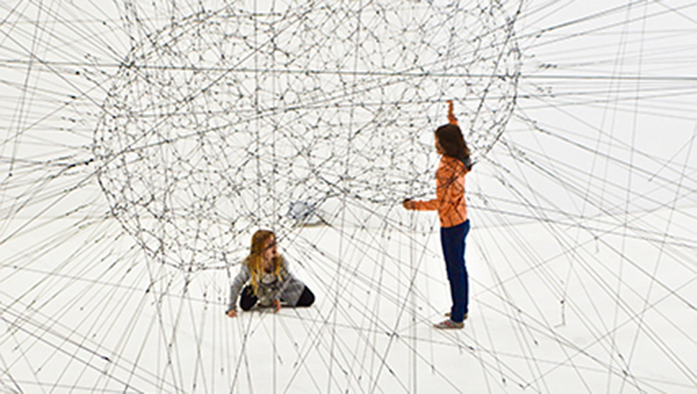Photographic Representation of AI (Artificial Intelligence) as peanut-shaped web of wires near two young women with crisscrossing wires branching off of center and filling the white room.