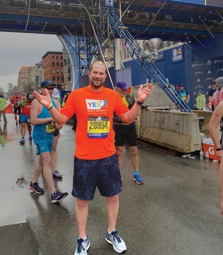 Runner in orange shirt with YES decal and Boston Marathon number reaches finish line and smiles at camera.